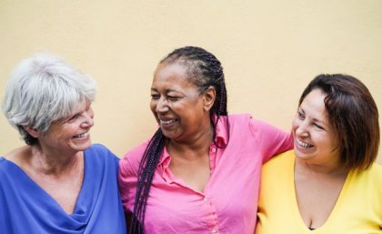 Three women in brightly coloured shirts smile and laugh. They have their arms around each other.